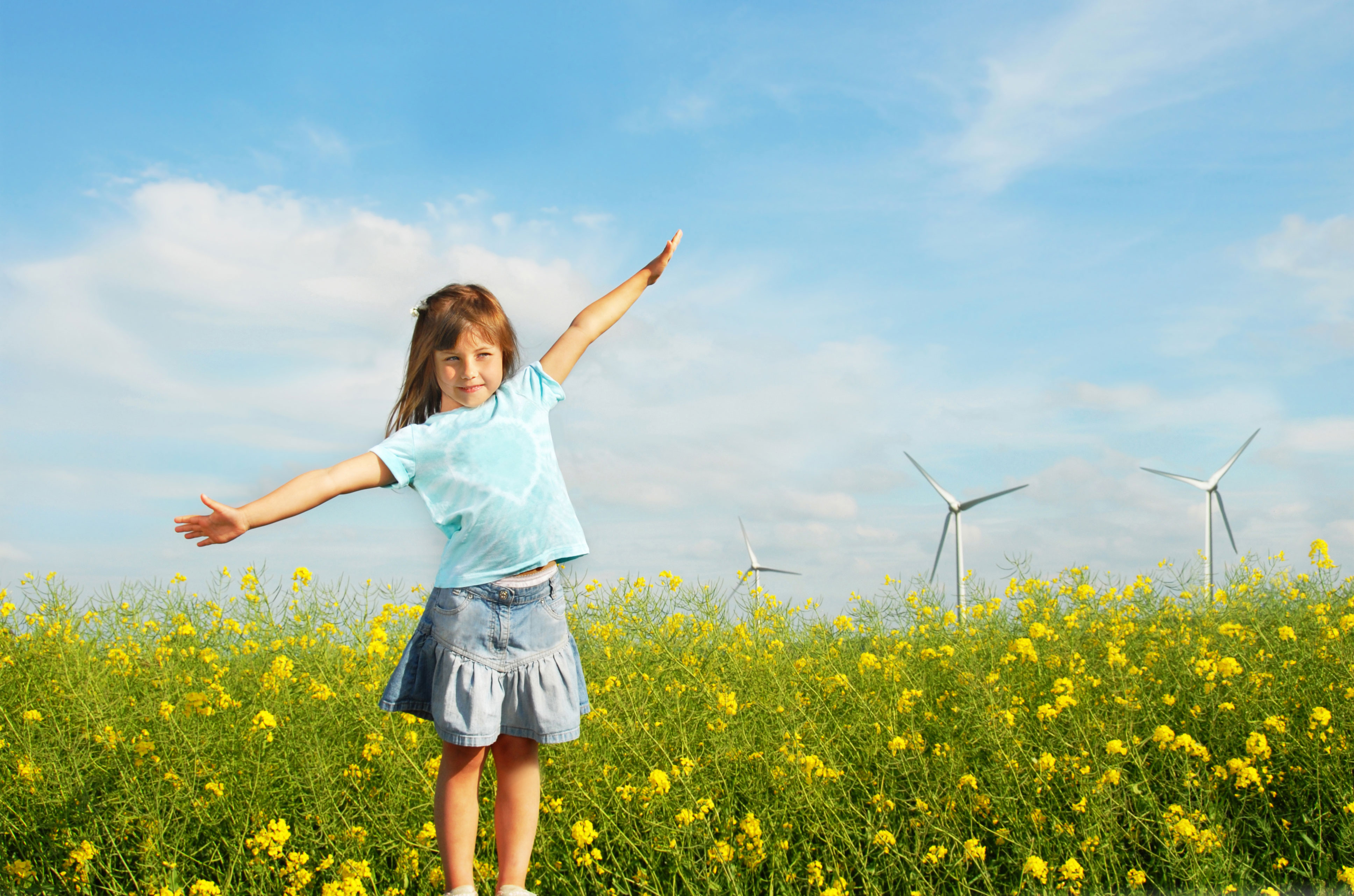 Flicka står med utsträckta armar i ett rapsfält med vindkraftverk bakom