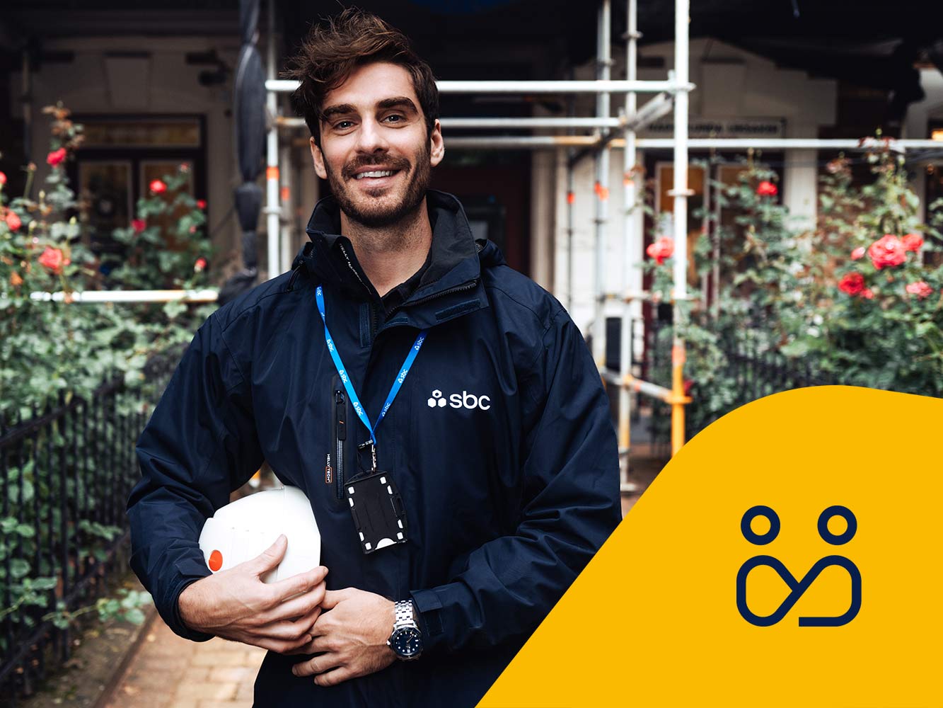 SBCs byggprojektledare Patrik står framför en fastighet med byggnadsställning och rosenrabatt. Projektledningsikon.