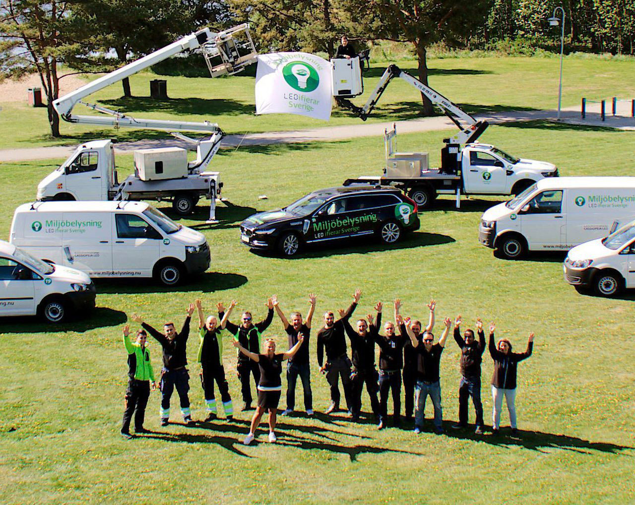 Stor grupp glada människor framför bilar på äng