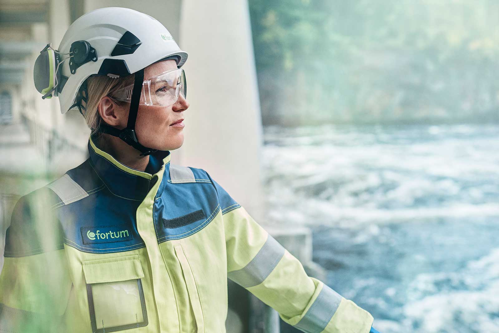 Kvinna med arbetskläder och skyddshjälm blickar ut över vattenfors