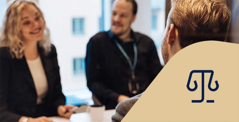 Tre jurister sitter vid ett bord och pratar och ler med ett anteckningsblock. Juridikikon.